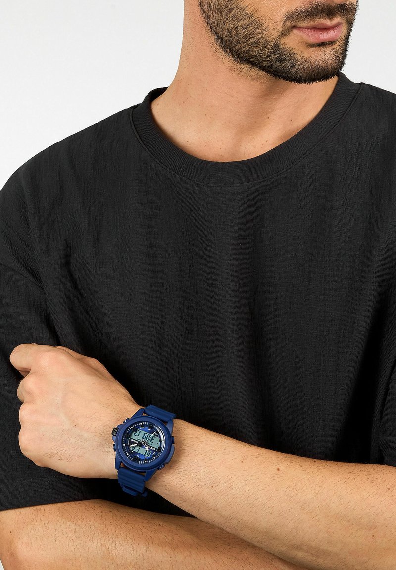 Orologio digitale blu con cinturino in gomma, dotato di un quadrante rotondo, display digitale e dettagli testurizzati sul cinturino. Indossato su un polso di carnagione chiara.