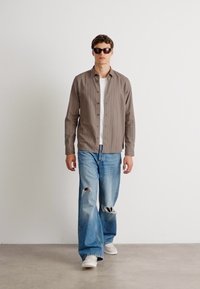 Camicia button-up a righe marroni sopra una t-shirt bianca, abbinata a jeans chiari strappati e sneakers bianche. Il modello indossa occhiali da sole neri.