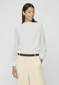 Blusa grigia chiara a maniche lunghe con un motivo a zigzag testurizzato, scollatura arricciata e polsini elasticizzati, abbinata a pantaloni beige a vita alta.