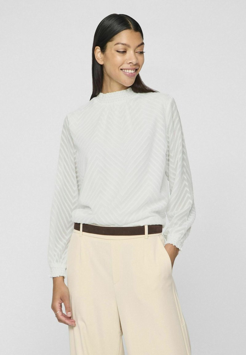 Blusa grigia chiara a maniche lunghe con un motivo a zigzag testurizzato, scollatura arricciata e polsini elasticizzati, abbinata a pantaloni beige a vita alta.