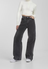 Valkoiset leveälahkeiset farkut mustalla denim-kankaalla, kuluneella tekstuurilla, korkealla vyötäröllä, etutaskuilla ja napilla kiinnitettävällä sulkijalla, yhdistettynä valkoiseen lyhytmalliseen paitaan.