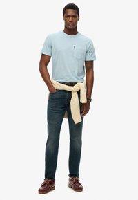 Camiseta de algodón azul claro con bolsillo; vaqueros azul oscuro; suéter crema atado a la cintura; zapatos de cuero marrón; reloj de plata en la muñeca.