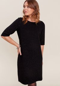 Robe noire en tricot avec un motif texturé, encolure ronde et manches trois-quarts. Présente des accents métalliques subtils.