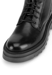 Melnas ādas šņorējamās zābaku ar biezām, tekstūrētām gumijas zole un sānu rāvējslēdzēju, rādot priekšpusi un ārējo sānu detaļas uz baltā fona.