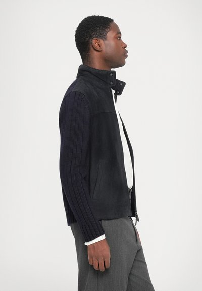 Veste marine avec un corps en daim et des manches en tricot, un col montant, une fermeture éclair et deux poches latérales. Texture lisse et design moderne.