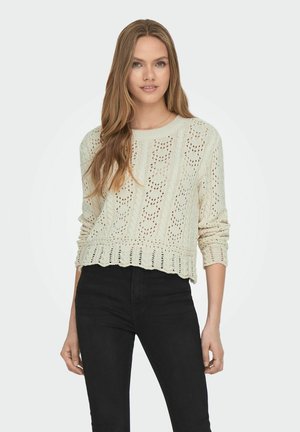 Hellbeige gestrickter Pullover mit lockerem Schnitt, der ein strukturiertes Muster und einen gerippten Saum aufweist, kombiniert mit schwarzen High-Waist-Hosen.
