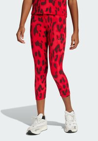 Leggings capri rojos con estampado de leopardo negro, con una cinturilla elástica y un logo bordado en el lateral, combinados con zapatillas blancas.