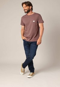 Een glimlachende man met een baard, gekleed in een mauve T-shirt met korte mouwen, donkerblauwe jeans en beige sneakers, staande met één hand in zijn zak tegen een beige achtergrond.