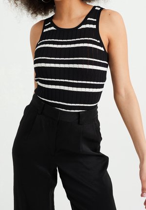 Femme portant un haut noir sans manches avec des rayures horizontales blanches et un pantalon noir taille haute sur fond blanc.