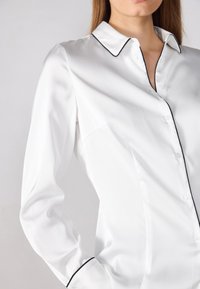Camicia in satin bianco con bordi neri, caratterizzata da un colletto classico, chiusura con bottoni e maniche lunghe con una texture liscia.