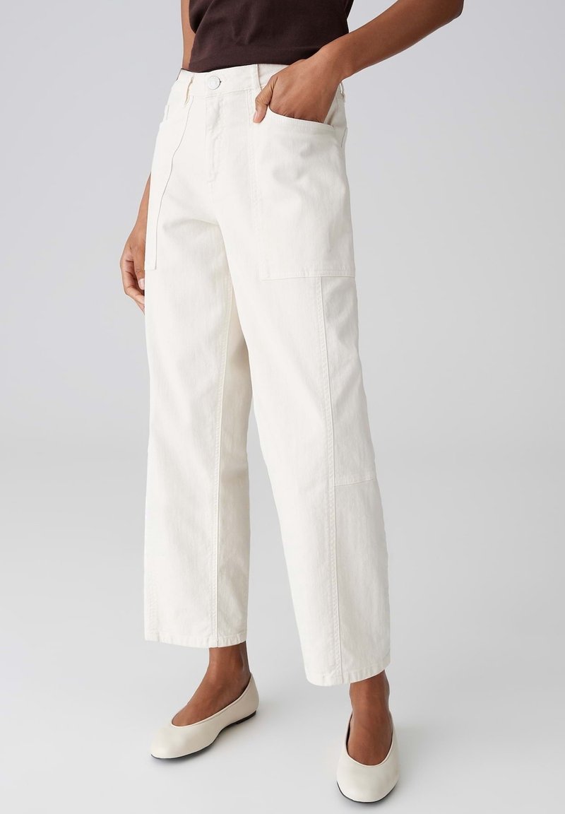 Personne portant un pantalon blanc à jambes larges avec des poches et des chaussures plates couleur crème, debout avec une main dans la poche.