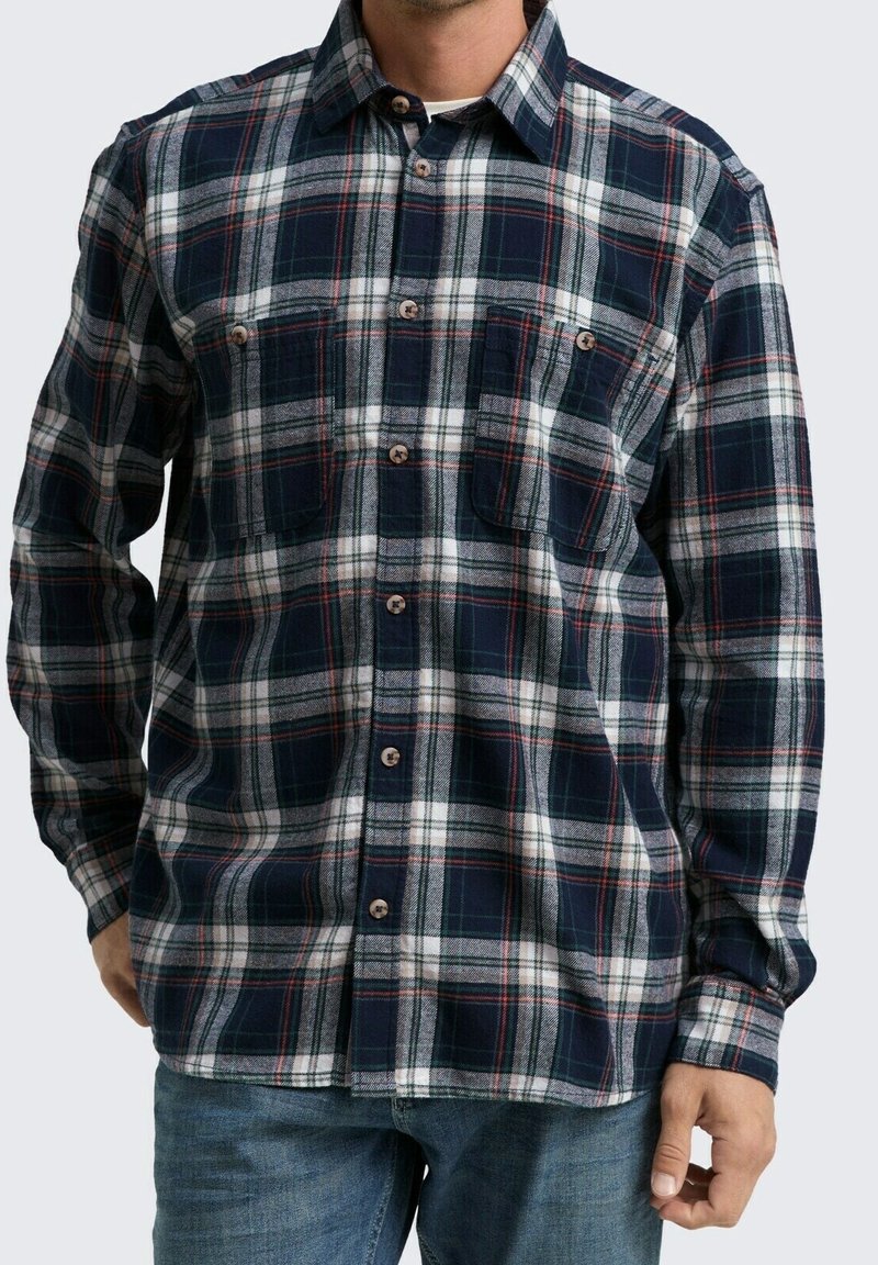 Homme portant une chemise à carreaux à manches longues boutonnée avec des lignes bleu marine, blanches et rouges, assortie à un jean bleu, debout avec une main dans la poche.