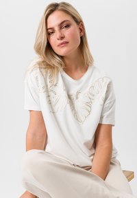 Witte t-shirt met gouden borduursels van bladeren op de voorkant, ronde hals, en korte mouwen, gecombineerd met lichtbeige broek.
