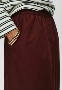 Pantalons larges bordeaux avec une taille élastique. Associés à un haut à rayures à manches longues. Texture lisse, poche latérale visible et bracelet en or.