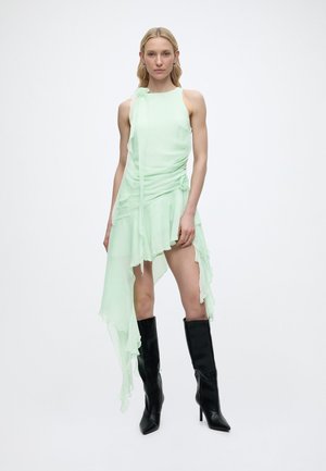 Γυναίκα που φοράει ανοιχτό πράσινο ασύμμετρο αμάνικο φόρεμα με βολάν και μαύρες μυτερές μπότες μέχρι το γόνατο, στέκεται μπροστά σε λευκό φόντο.