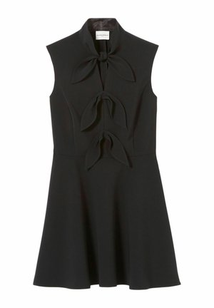Mujer joven que lleva un vestido negro sin mangas con lazos, sandalias negras con tiras, bolso negro y un accesorio en su brazo superior, de pie frente a un fondo sencillo.