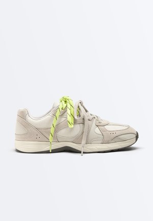 Beige athletic shoe with light green and beige mismatched laces, textured sole, and suede accents on a white background.
