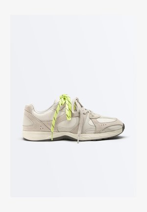 Beige sportssko med lysegrønne og beige mismatchede snørebånd, tekstureret sål og ruskindsdetaljer på hvid baggrund.