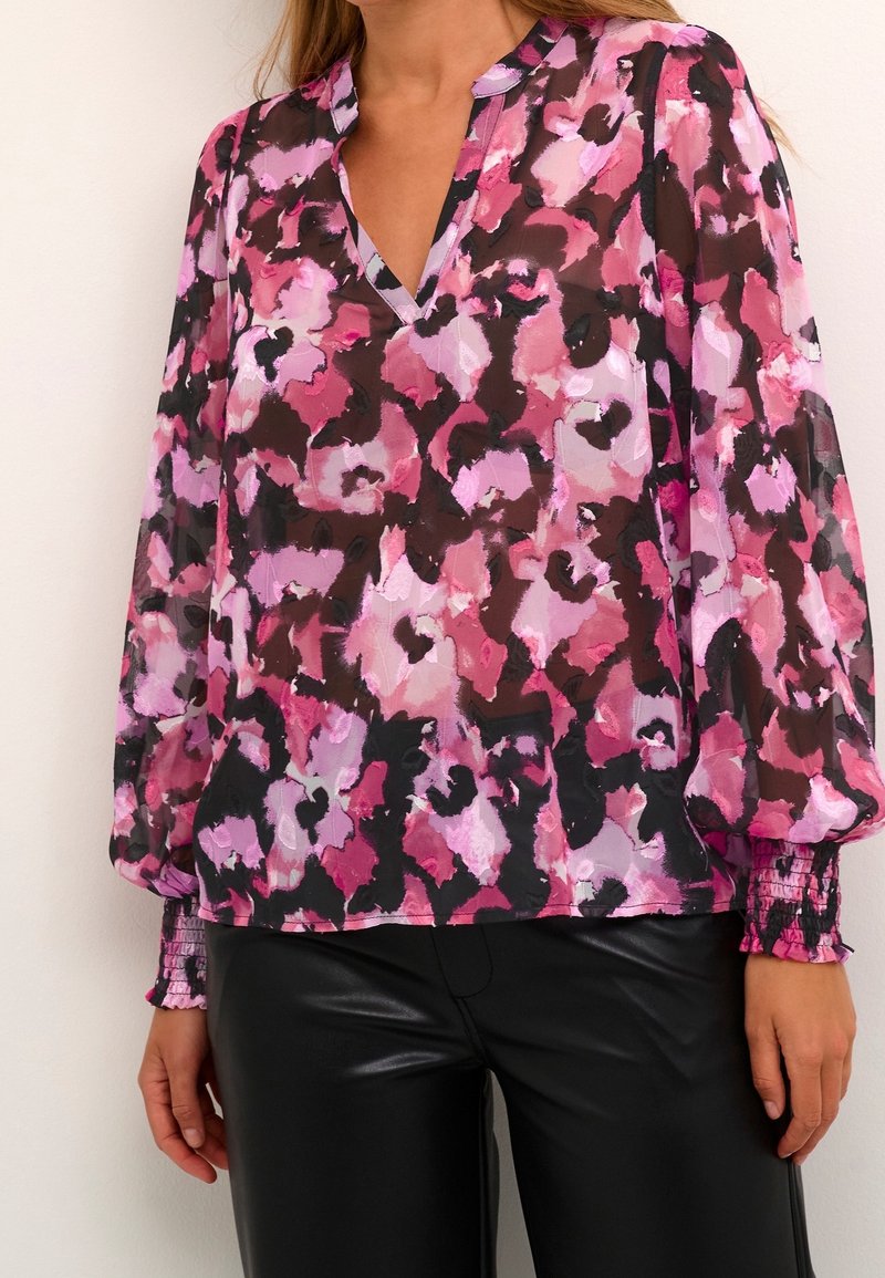 Kvindelig person iført gennemsigtig bluse med lange ærmer i pink, lilla og sort blomsterprint samt sorte læderbukser, som står op ad en lys væg.