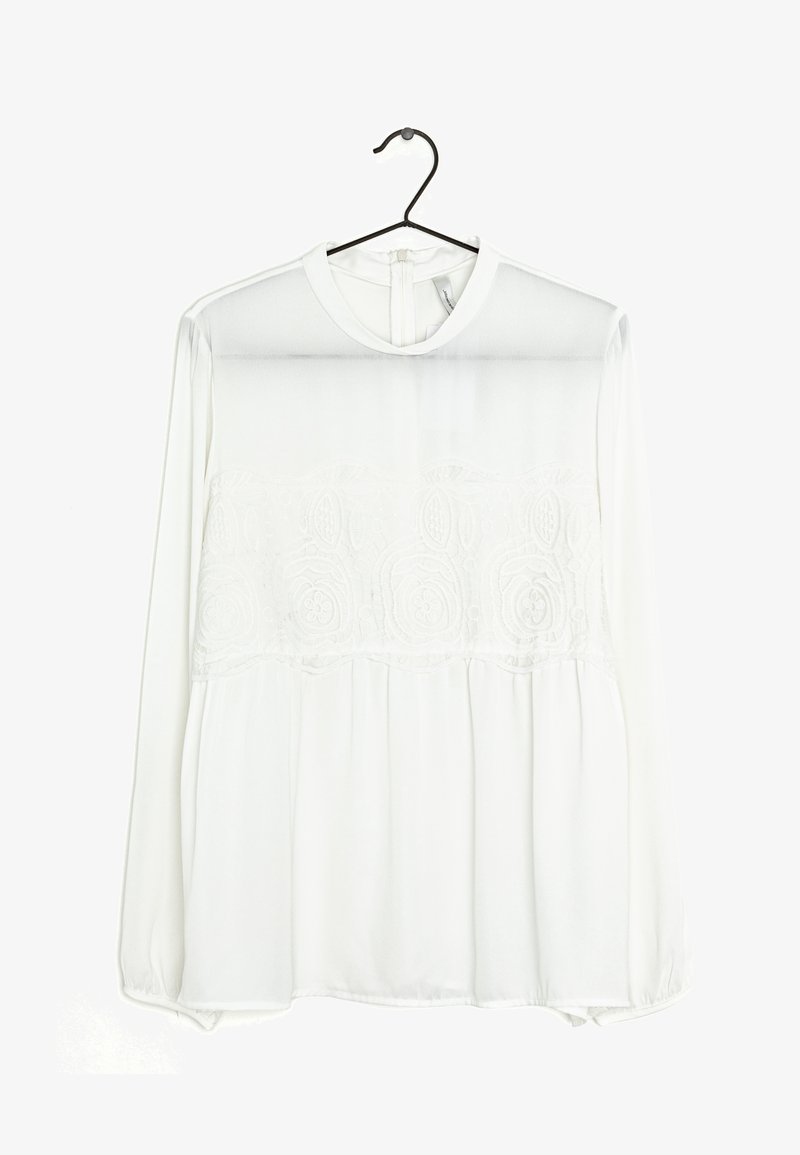Blouse blanche à manches longues avec un détail en dentelle florale sur la poitrine, col rond et fermeture boutonnée au dos, suspendue sur un cintre noir.