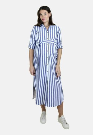 Gravid kvinde iført en skjortekjole med blå og hvide lodrette striber, der bindes i taljen, og hvide sneakers, stående mod en ensfarvet baggrund.