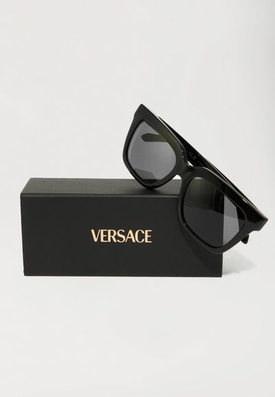 Černé oversized sluneční brýle s tmavými skly, se silným rámem a ostrým designem. Dodávají se v černé krabičce s nápisem "VERSACE" v zlaté barvě.
