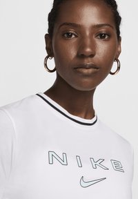 Fehér póló fekete és kék Nike logóval, bordázott nyakkivágással fekete kiegészítőkkel. A modellen fülbevalók vannak.
