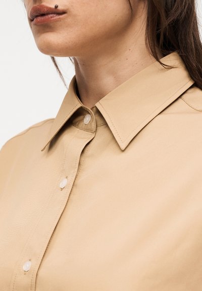 Κλείσιμο ενός ατόμου που φοράει μπεζ πουκάμισο με κουμπιά και μυτερό γιακά, δείχνοντας το κάτω μέρος του προσώπου και τον λαιμό.