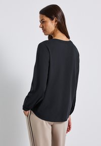 Una donna con capelli castani lisci indossa una blouse nera a maniche lunghe e pantaloni beige con strisce laterali nere, in piedi davanti a uno sfondo chiaro e uniforme.