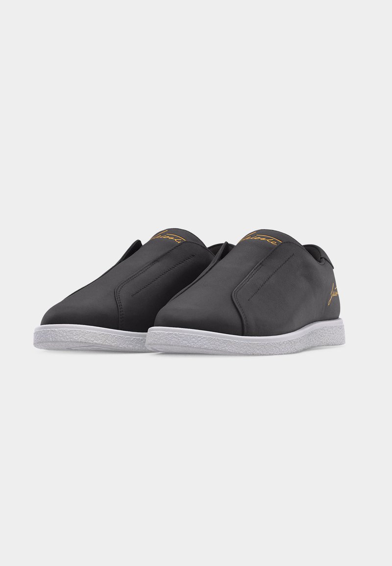 Coppia di sneakers nere slip-on con suole bianche testurizzate e discreto marchio dorato sulla linguetta e sul lato, posizionate su uno sfondo chiaro.