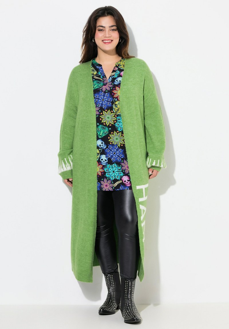 Largo cárdigan verde con mangas amplias y detalle de flecos. Debajo, una camiseta negra con patrones coloridos de flores y calaveras. Leggings negros y botas con tachuelas.