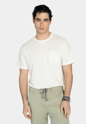 Joven con camiseta blanca de manga corta con bolsillo y pantalones gris claro con cordón, expresión neutral, de pie frente a un fondo liso.