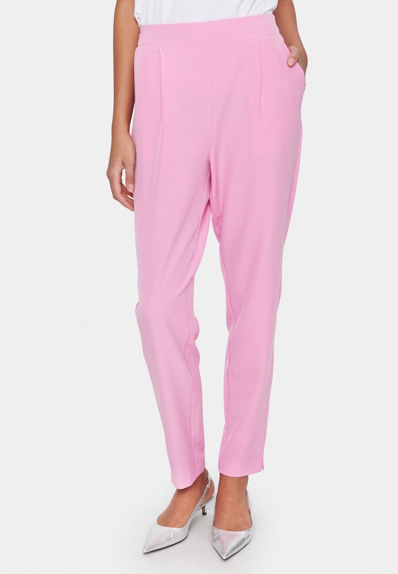 Femme portant un pantalon fuselé rose pâle avec des poches latérales, associé à des escarpins pointus argentés, debout devant un fond uni.
