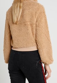 Beżowa kurtka ze sztucznego futra o krótkim kroju i ściągaczu na dole. W połączeniu z czarnymi, wysokim stanem jeansami z czerwonymi paskami po bokach.