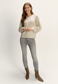 Béžový pletený sveter s geometrickými vzormi, rebrovaným lemom a balónovými rukávmi, spojený so svetlošedými džínsami a taupe členkovými čižmami.