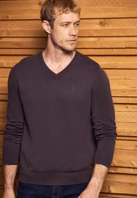 Mörklila v-ringad tröja i mjuk stickad kvalitet, med långa ärmar och ribbade muddarna och fållen. Enkel logotypdetalj på bröstet.