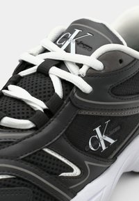 Μαύρα αθλητικά παπούτσια με επάνω μέρος από δίχτυ και συνθετικές δερμάτινες λεπτομέρειες, λευκές κορδόνες και μια εξέχουσα ασημένια λογότυπο στη γλώσσα.