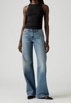 Kobieta ubrana w czarny top bez rękawów, wysokiej talii wyblakłe szerokie niebieskie dżinsy i czarne buty z ostrym noskiem, stojąca na jednolitym tle.