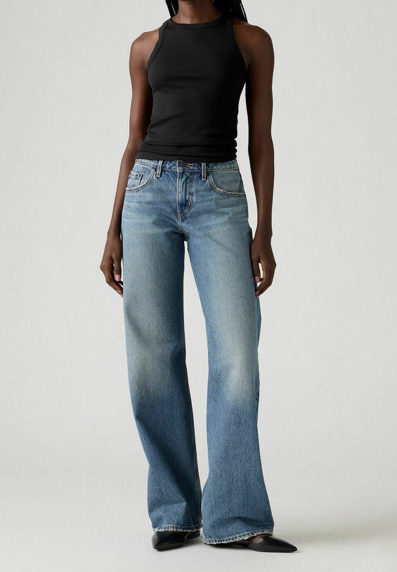 Femme portant un haut noir sans manches, un jean large bleu délavé taille haute, et des chaussures noires à bout pointu, debout devant un fond uni.