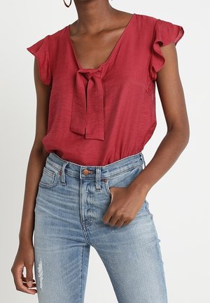 Blusa rossa con dettaglio a fiocco al collo, maniche corte a volant, abbinata a jeans chiari a vita alta con effetti di usura.