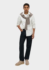 Camisa blanca a rayas, suéter gris atado alrededor de los hombros, pantalones de mezclilla oscuros, zapato beige. Modelo de pie con la mano en el bolsillo, fondo neutro.