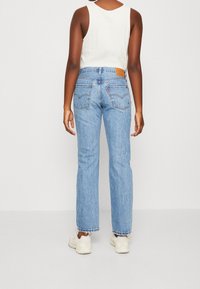 Jeans de mezclilla azul claro con pierna recta, dobladillo deshilachado y bolsillos traseros con un parche de cuero. Llevados con una blusa sin mangas blanca.