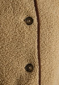 Beež fliisriie, millel on tekstuuriga, plüüšine pind. Omab pruuni kanga ääre ning ümmargust metallnuppu, millel on kiri "JOULES EST. 1990".