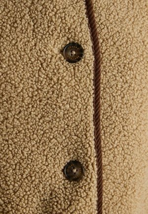 Beige fleece fabric with a textured, plush surface. Features a brown corduroy trim and a round metal button with “JOULES EST. 1990” inscription.