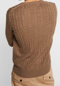 Pull en maille marron avec un motif en câble, présentant un col rond et des poignets et un ourlet côtelés. Texture et élasticité visibles dans le tissu.
