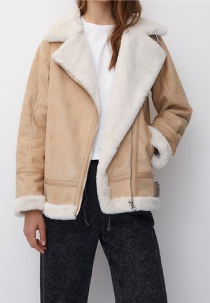 Personne portant une veste en peau lainée beige avec bordure en fourrure blanche, sur une chemise blanche et un pantalon noir, debout devant un fond uni.
