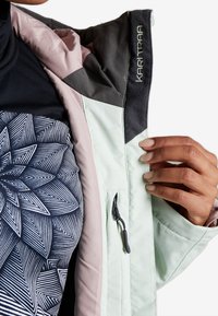 Miętowa zielona kurtka z szarymi i różowymi akcentami, wyposażona w kieszeń na suwak, miękką teksturę oraz wewnętrzną wzorzystą bluzkę z geometrycznymi liniami.