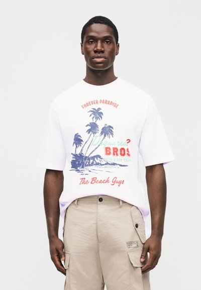 Mann trägt weißes T-Shirt mit Palmenmotiv und dem Text "Forever Paradise", kombiniert mit beigen Cargo-Shorts vor einfarbigem Hintergrund.