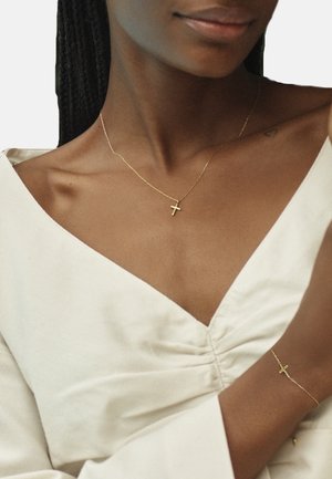Goldfarbener Kreuzanhänger an einer zarten Kette, getragen mit einer weißen Bluse im V-Ausschnitt, was eine minimalistische Ästhetik unterstreicht.