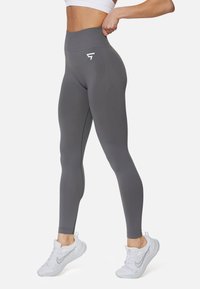 Hoch taillierte graue Leggings mit glatter Textur, ausgestattet mit einem weißen Logo an der Hüfte und figurbetontem Design, kombiniert mit weißen Sportschuhen.
